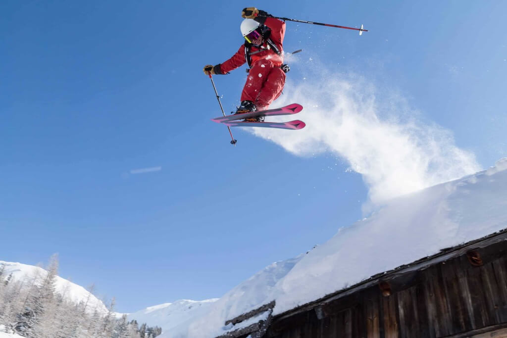 Skifahrer springt über eine schneebedeckte Hütte in den Alpen bei strahlendem Himmel.
