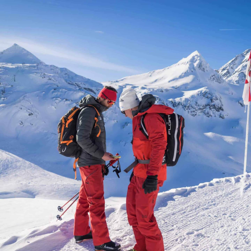 Zwei Skibergsteiger überprüfen die Ausrüstung vor beeindruckender Alpenkulisse.