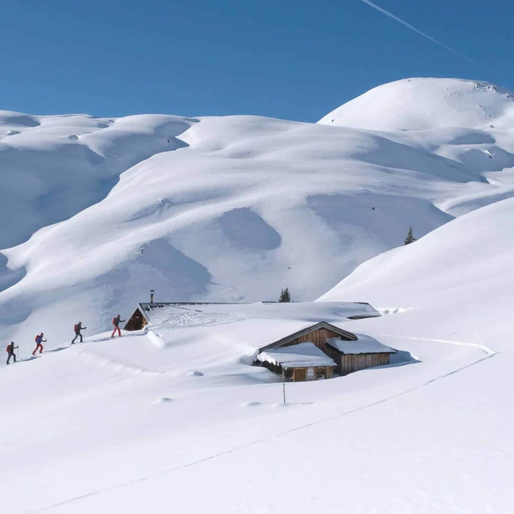 Skifahrer wandern durch verschneite Alpenlandschaft zu einer Berghütte.