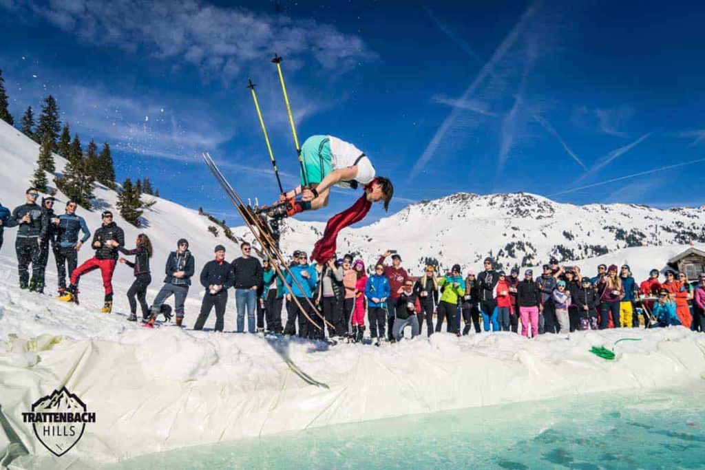 Skifahrer springt über ein Wasserbecken, Publikum jubelt im schneebedeckten Gelände.