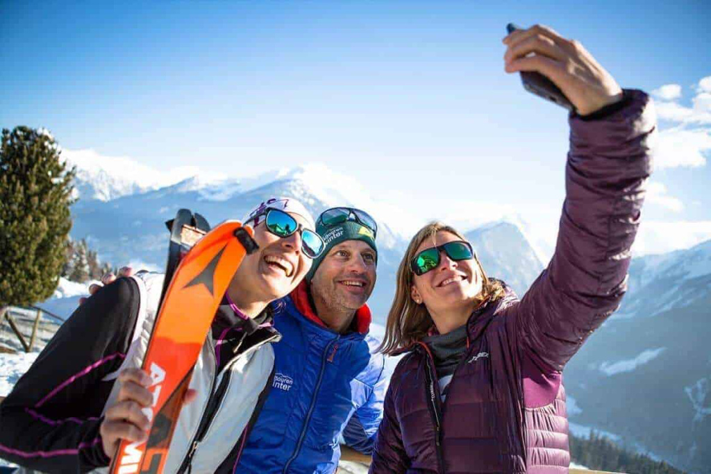 Drei Skitouren-Teilnehmer machen ein Selfie vor alpiner Landschaft im Winter.