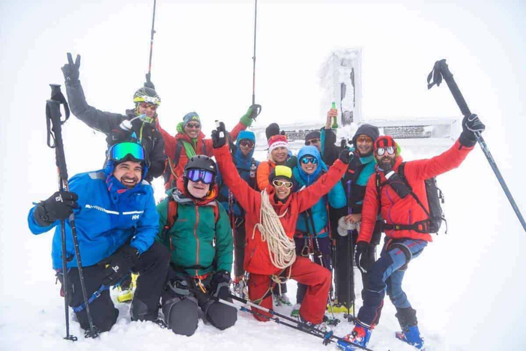 Gruppe von Skibergsteigern feiert am Gipfel, umgeben von Schnee und einem Kreuz.