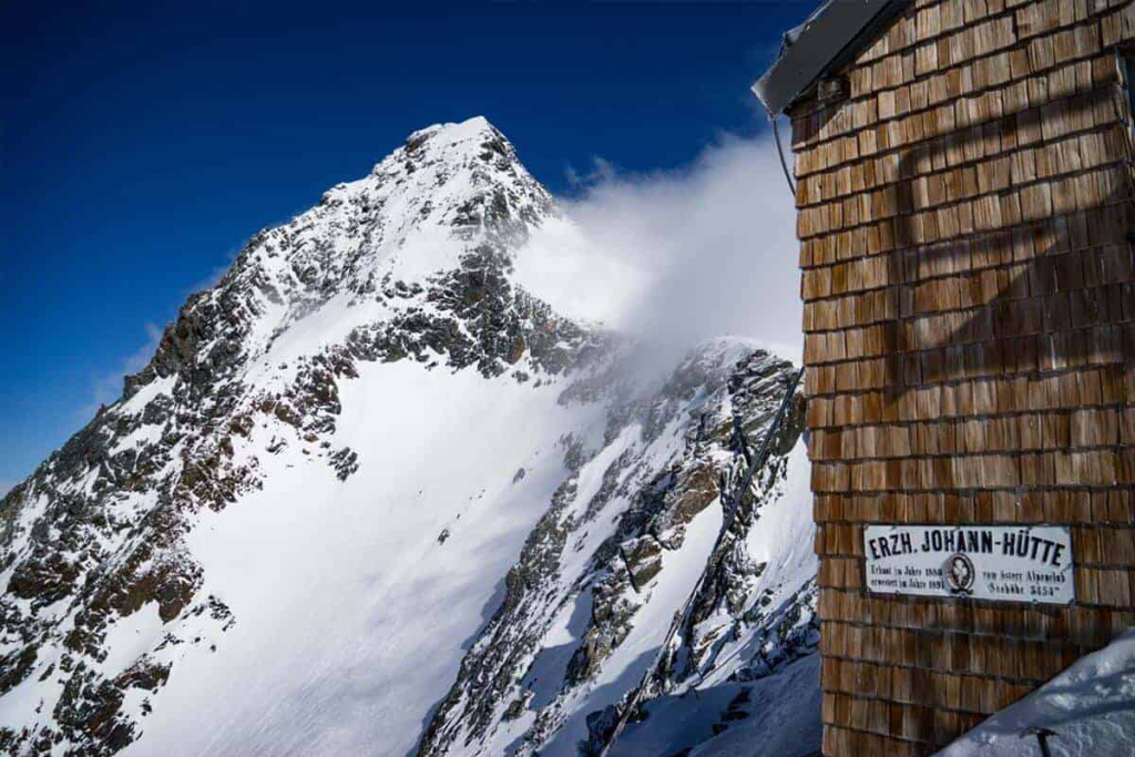Erzh. Johann-Hütte mit schneebedecktem Großglockner im Hintergrund, strahlend blauer Himmel.