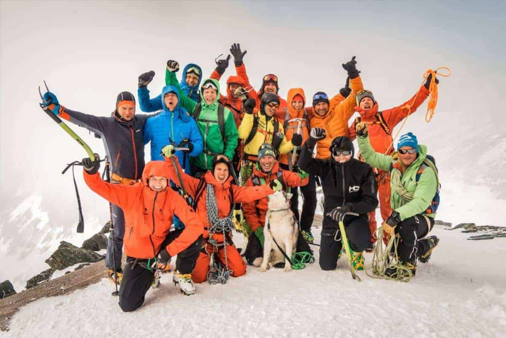 Gruppe von Skitourengehern feiert am Gipfel in bunten Outfits mit einem Hund.
