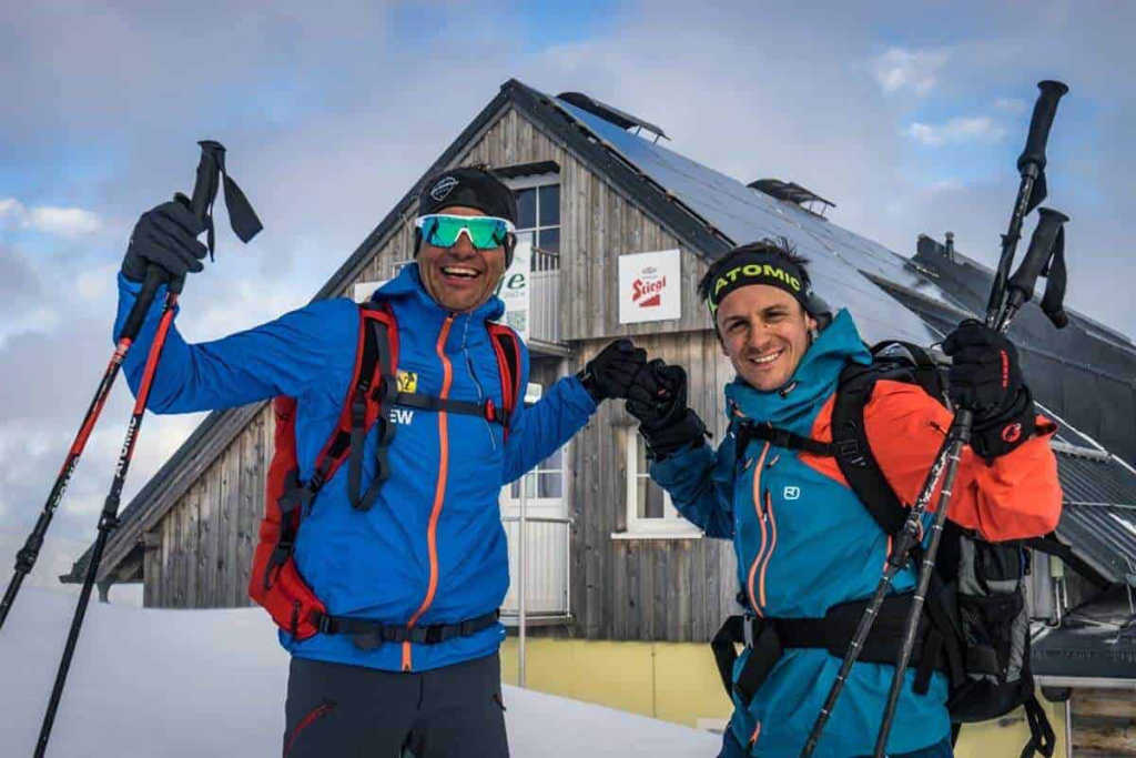 Bergführer feiern die Erreichung eines Gipfels vor einer Berghütte im Schnee.