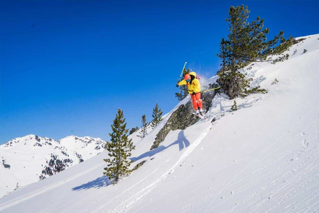 Skitourenfahrer springt über Schnee mit Blick auf die Alpen unter strahlend blauem Himmel.