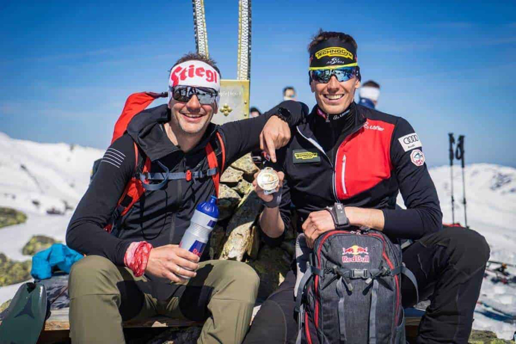 Zwei Skifahrer posieren auf einem Gipfel in den Alpen, umgeben von Schnee und Berglandschaft.