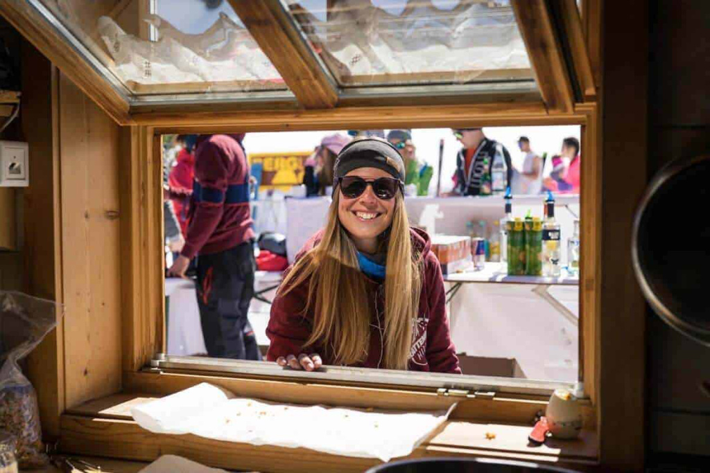 Fröhliche Frau am Fenster mit Sonnenbrille, im Hintergrund Skifahrer und Getränke.
