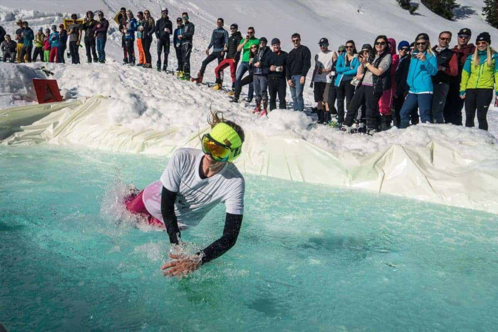 Skifahrerin springt in ein Wasserbecken, umgeben von zuschauenden Skifans im Schnee.