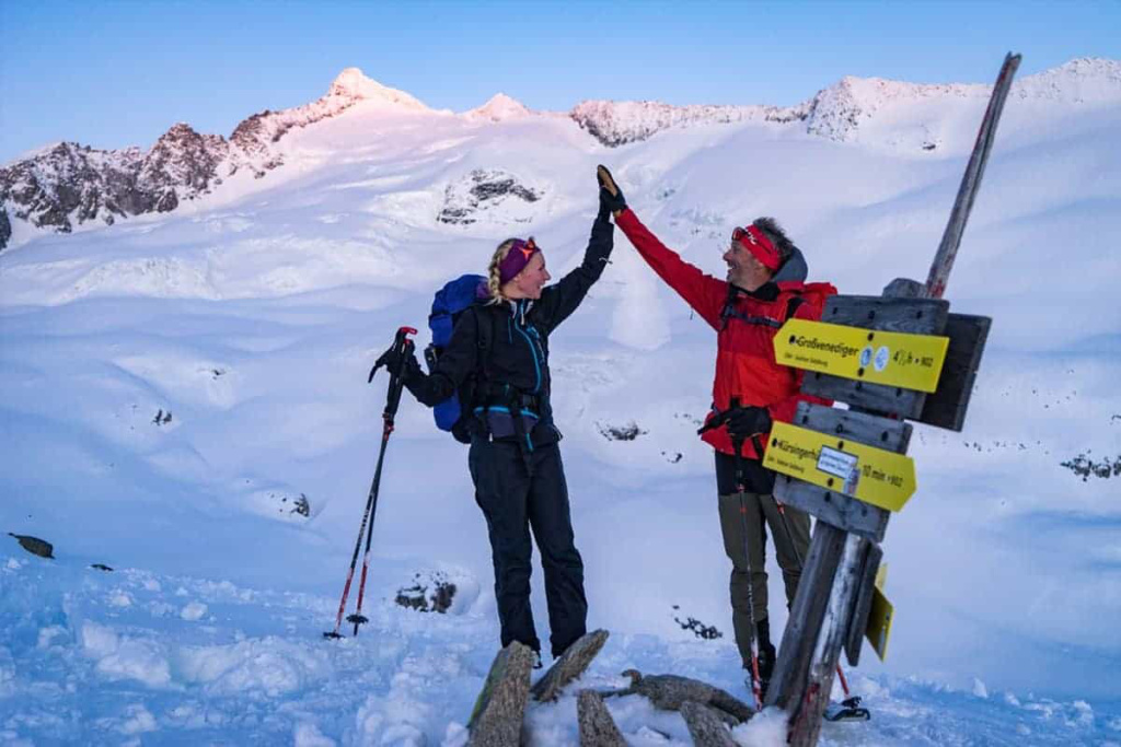 Zwei Skitourengeher feiern am Gipfel mit den Alpen im Hintergrund.