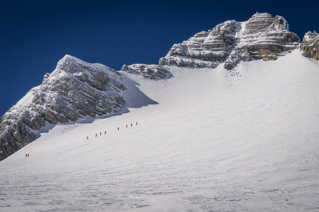 Skitouren-Gruppe besteigt schneebedeckten Hang in den Alpen unter blauem Himmel.