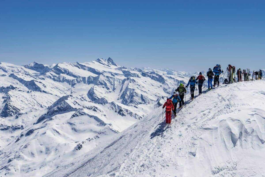 Skibergsteiger auf einem verschneiten Gipfel mit Alpenpanorama im Hintergrund.