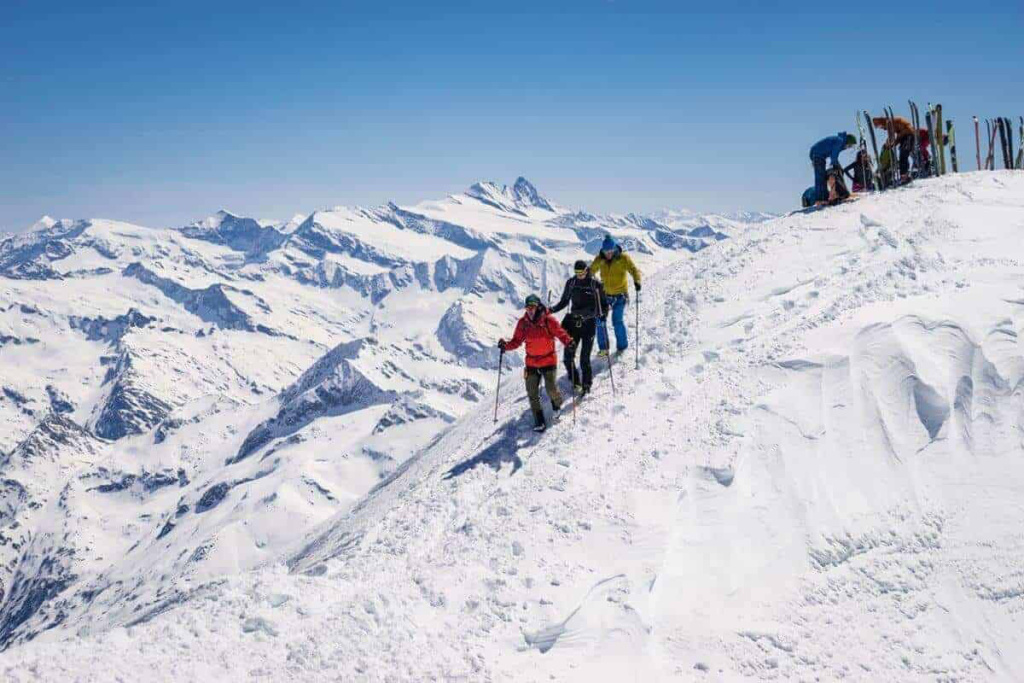Skitourengeher auf schneebedecktem Gipfel mit Alpenpanorama im Hintergrund.