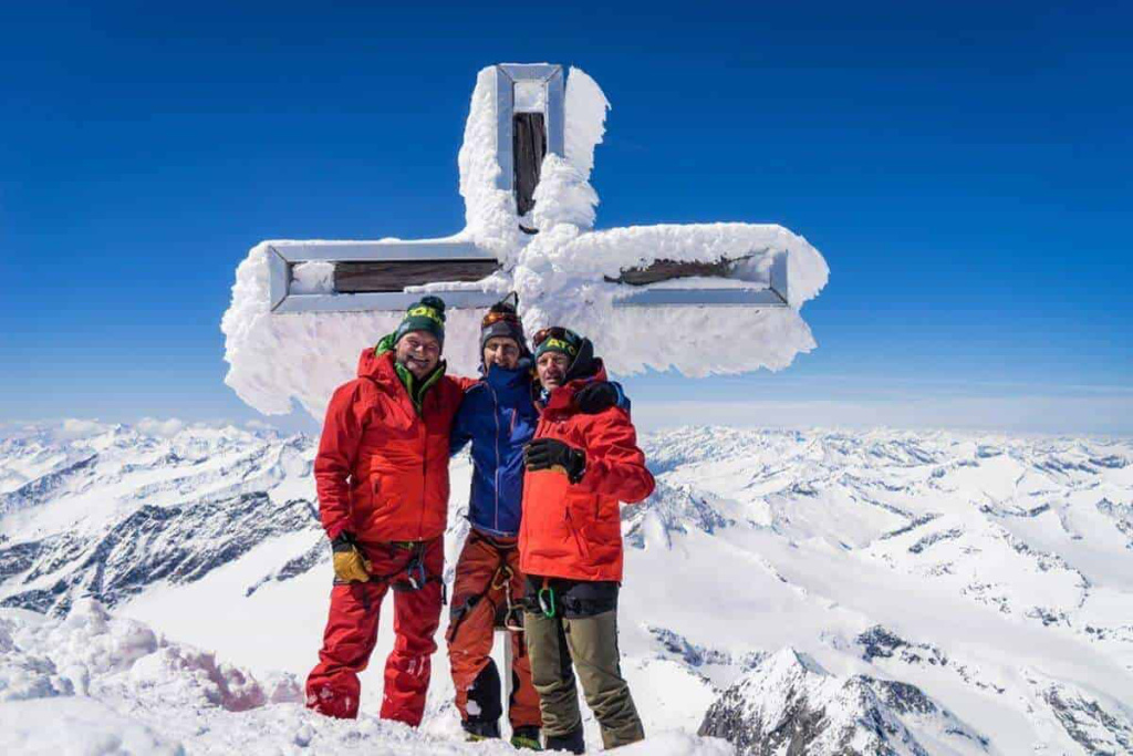 Bergsteiger in rot-orange Skibekleidung posieren am schneebedeckten Gipfel mit Kreuz.