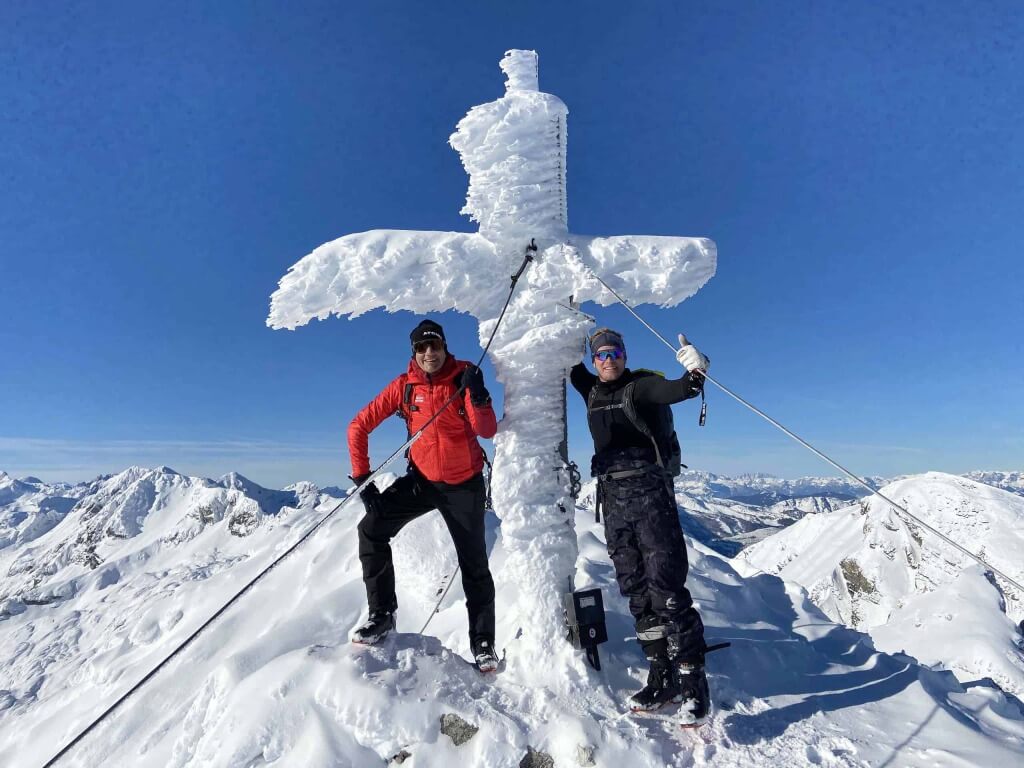 Zwei Skitourengeher posieren am schneebedeckten Gipfel mit einem gefrorenen Kreuz im Hintergrund.