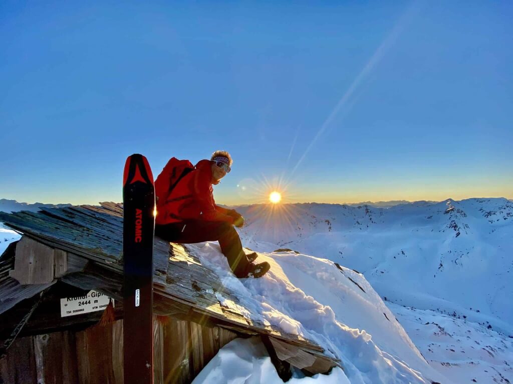 Skitourengeher auf schneebedecktem Dach, im Hintergrund strahlender Sonnenaufgang über den Alpen.