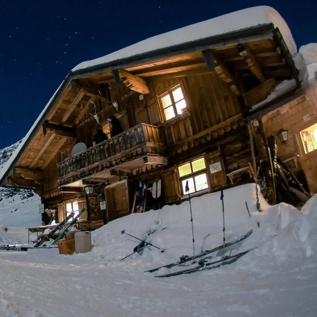Gemütliche Berghütte im Schnee bei Nacht, umgeben von alpiner Winterlandschaft.